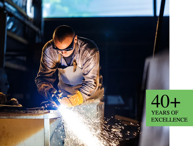 A welder wearing protective glasses and a leather apron uses a torch, showering bright sparks downward in a workshop.